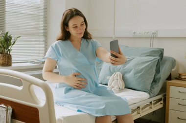 Genç müstakbel anne, doğum hastanesinde yatağında otururken içtenlikle gülümsüyor ve selfie çekiyor.