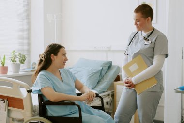 Young nurse holding yellow folder and listening to young woman in labor while she sitting in wheelchair clipart