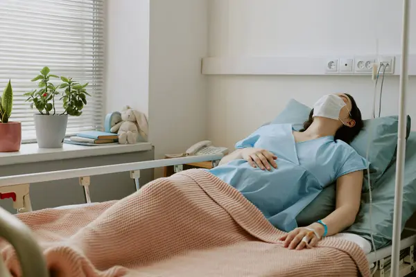 stock image Pregnant patient with medical mask on face sleeping on bed and holding her hand on belly while she under dripper