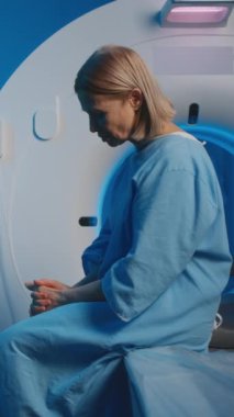 MRI odasındaki tarama yatağında oturan, radyoloji hemşiresinin tanısal prosedürü izlemesini ve kontrol etmesini bekleyen mavi hastane önlüklü, orta boy, beyaz kadın dikey görüntüsü.