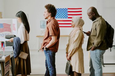 Oy kullanma merkezinden oy pusulası almak için sırada bekleyen etnik çeşitliliğe sahip gençlerin orta uzunluktaki yan görüntüsü