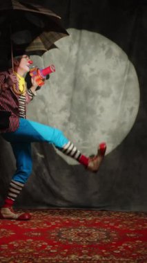 Olgun beyaz erkek sirk sanatçısının sahnede komik çizimler gösterip el yapımı oyuncak silah ve şemsiyeyle oynadığı dikey bir sahne.