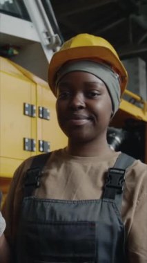 Şapkalı ve iş kıyafetli genç siyah kadının dikey portresi. Kameraya bakıyor ve endüstriyel fabrikada çalışırken gülümsüyor.