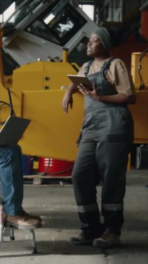 İş kıyafetleri içindeki genç siyahi kadın teknisyenin iş günü iş yerinde iş arkadaşlarıyla sohbet ederken, gülümserken ve dijital tablet kullanırken tam bir dikey çekimi.