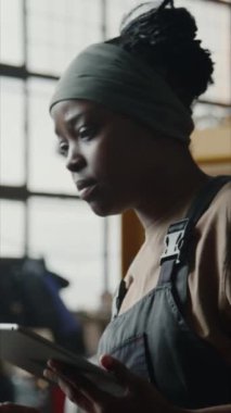 Genç Afrikalı Amerikalı bir kadının iş kıyafetlerini dikey çekimde fabrika ekipmanlarını kontrol ederken ve dijital tablet üzerinde yazarken.