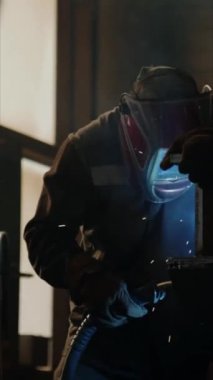 Yüz kalkanı ve iş kıyafetindeki kaynakçının dikey görüntüsü endüstriyel fabrikada kaynak makinesiyle metal işi yapıyor.