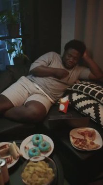 Şişman siyahi bir adamın kanepenin yanındaki fast food 'da oturup TV seyredip akşam patates kızartması yediği dikey bir sahne.