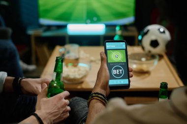 Tanımlanamayan bir adam arkadaşlarıyla TV 'de futbol maçı izleyip akıllı telefondan en sevdiği takıma bahis oynuyor.
