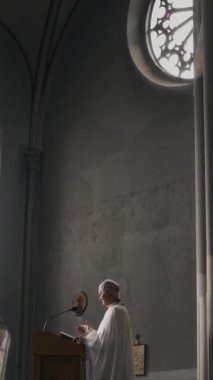Beyaz cüppeli kıdemli papazın mikrofonla podyumda durup gotik kilisede İncil 'le vaaz verirken dikey olarak aşağı eğilmesi.