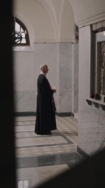 Siyah cüppeli rahibin dikey resmi mermer döşemeli kilisede elinde yanan bir mum tutarak dua ederken.
