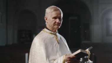 Beyaz şezlonglu yaşlı beyaz saçlı papazın portresi. Elinde İncil, kilisede kameraya poz veriyor.