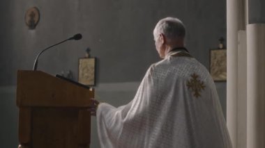 Beyaz cüppeli kıdemli papaz elinde mikrofonla podyuma çıkıyor ve kilisede vaaza hazırlanırken İncil 'i açıyor.