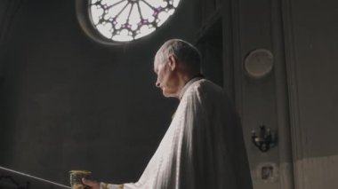 Yaşlı rahip, loş aydınlatılmış kilisede kutsal tören sırasında elinde süs kadehiyle uzun camdan içeri ışık süzülürken...