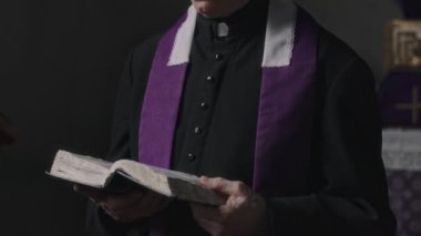 Kıdemli rahibin siyah cüppeli, mor atkılı, İncil okuyan ve loş bir kilisede kameraya bakan portre fotoğrafını kaldır.