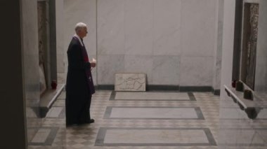 Siyah bornoz ve mor şal giymiş, elinde mumla mermer döşeli kilisede yürüyen, mihrabın önünde durup Tanrı 'ya dua eden kıdemli rahiplerle dolu bir fotoğraf.