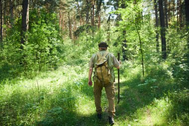 Sırt çantası takmış, tahta sopayla orman boyunca yürüyüp, uzayı kopyalayan, tanınmayacak kadar olgun bir adamın arka görüntüsü.