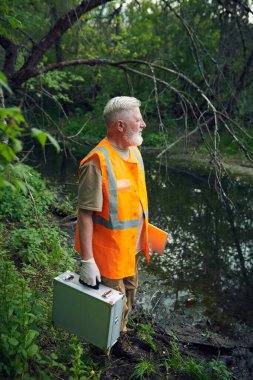 Vertical high angle view long shot of senior ecologist holding portable lab kit standing at bank of river in forest clipart