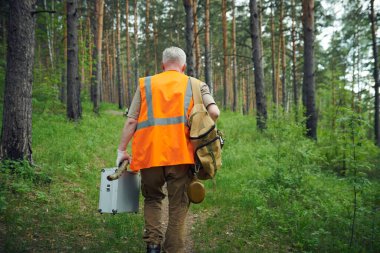 Güvenlik yeleği giymiş, elinde test seti valiziyle dağ ormanlarında yürüyen tanınmayan kıdemli bir ekolojistin arkadan çekilmiş fotoğrafı.