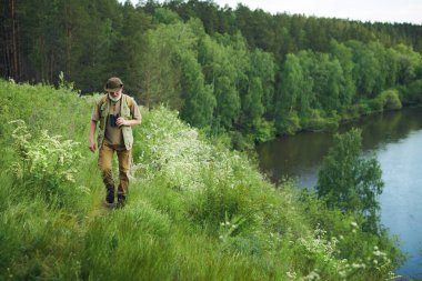 Wide shot of senior Caucasian man wearing backpack hiking along bank of mountain river on summer day, copy space clipart