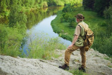 Dağ nehrinin kıyısındaki kayanın üzerinde sırt çantası takan tanınmayan yetişkin doğa bilimcilerin geniş açılı görüntüsü.