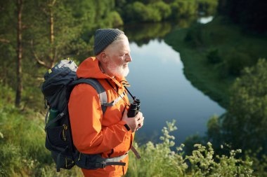 Turuncu ceketli, bereli ve sırt çantalı üst düzey beyaz erkek turist nehir kenarında yürüyüş yapıyor.