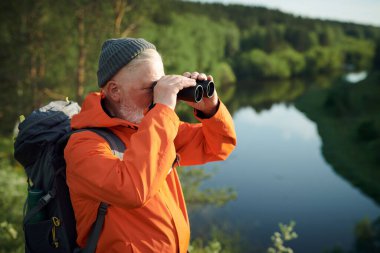 Dürbünle nehir kıyısında duran sırt çantalı, tanınmayan yetişkin erkek turistin orta boy fotoğrafı.