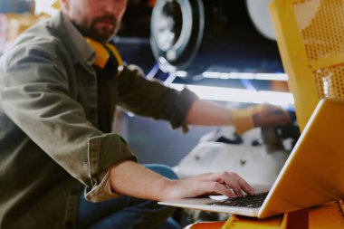 İşyerinde endüstriyel makinenin parçalarını tamir ederken bilgisayarını kullanan sakallı tamircinin seçici odak noktası.