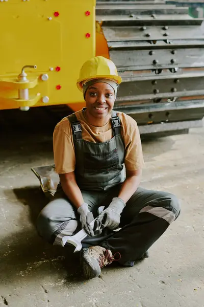 stock image Vertical shot of happy young African American female technician wearing hardhat sitting on floor with wrench in hands smiling at camera