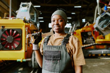 Fabrikadaki iş yerinde kameraya poz veren neşeli Afro-Amerikan kadın teknisyenin orta boy portresi.