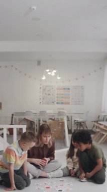 Kafkas kadın eğitmenin, anaokulunda yerde oturup, çok ırklı anaokulu çocuklarıyla kart oyunu oynaması.