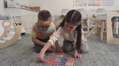 Kreş öncesi neşeli beyaz erkek ve kız halı döşemesinde oturup kart oynayarak birbirlerine beşlik çakarken arka planda pastel ahşap anaokulu içleri.