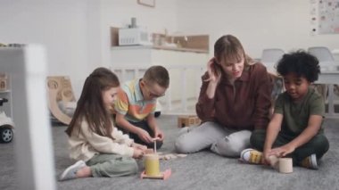 Anaokulunda yerde oturup ahşap oyuncaklar oynayıp rekabet eden bir grup anaokulu çocuğu. Beyaz kadın eğitmen elini sıkarak kazananı kutluyor.