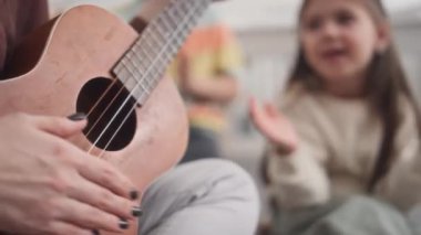 Gitar çalan tanınmayan bayan müzik öğretmeninin, anaokulunda okuyan ve coşkuyla el çırpan kız ve erkek çocukların ortasında.