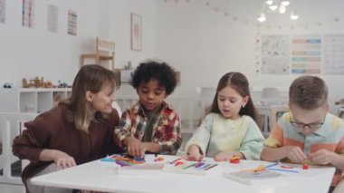 Beyaz kadın eğitimci modellik sürecini incelerken ve çocuklara övgüler yağdırırken, üç farklı çocuğun anaokulunda masalarda oturup renkli plastiklerinden figürler yaratırken orta boy bir fotoğraf.