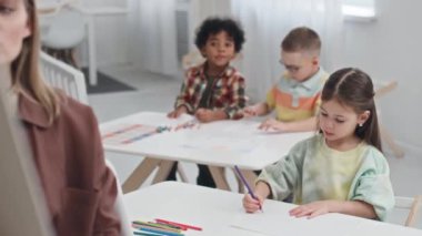 Anaokulunda çizim dersi sırasında çoklu etnik çeşitliliğe sahip çocuklara resim çizme tekniklerini açıklayan beyaz kadın resim öğretmeni.