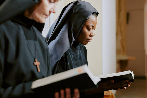 Selective focus medium closeup of two ethnically diverse nuns holding books in hands and reading religious texts