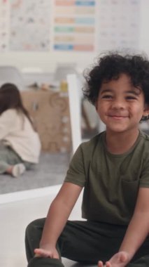 Sınıfta yerde oturmuş gülümseyen ve kameraya bakan kıvırcık saçlı neşeli Afrikalı Amerikalı çocuğun dikey portresi.