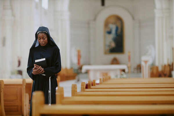 Medium long shot of young African American nun holding Bible book walking along nave in Catholic church, copy space