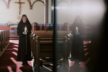 Kafkas Katolik rahibesinin elinde mum tutarken nave 'de durduğu ve kapı yüzeyindeki donuk yansımasının geniş bir görüntüsü.