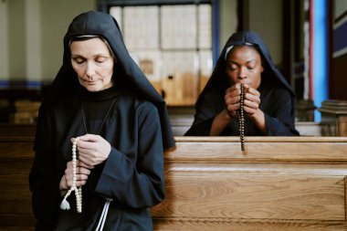 Kilisedeki sıralarda oturan ve ellerinde tespihle dua eden iki farklı etnik çeşitliliğe sahip genç ve olgun Katolik rahibe.