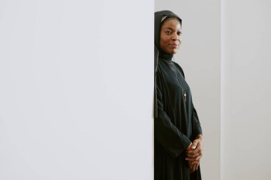 Beyaz duvarın arkasında duran kendinden emin genç Afrikalı Katolik rahibenin minimalist portresi.