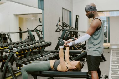 Afrika kökenli Amerikalı koç, kadın müşterisine kas yığını konusunda yardım ediyor. Birlikte göğüs presi yapıyorlar.