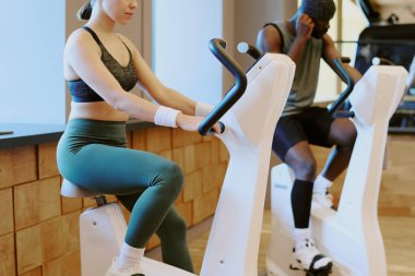 Orta boy, kaslı bir kadın ve egzersiz bisikletinde ısınan Afrikalı Amerikalı bir adam, kulaklığını ayarlayan bir adam.