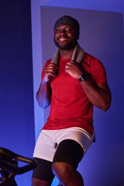 Afrikalı Amerikalı bir adam kardiyo treninden sonra dinleniyor ve terlemek için gri havlusunu tutuyor. Gülümsüyor ve içtenlikle dişlerini gösteriyor.
