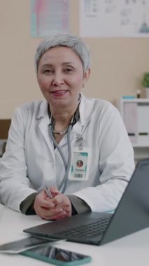 Kendine güvenen, beyaz önlüklü beyaz kadın jinekoloğun dikey portresi klinikte oturuyor, gülümsüyor ve kameraya bakıyor.