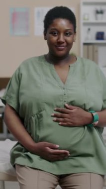 Klinik ortamda poz veren, gülümseyen ve gururla kameraya bakan hamile, Afro-Amerikan kadının dikey portresi.