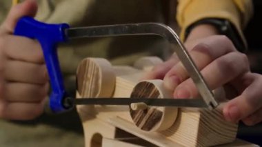 Ahşap işçiliği dersi sırasında tahta oyuncak tekerlekleri kesmek için demir testeresi kullanan çocuğun yakın çekim görüntüleri.