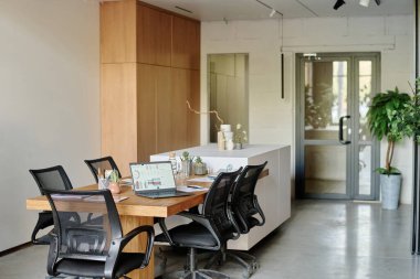 Ahşap masa ve siyah sandalyelerle donatılmış geniş ve aydınlık ofis, dijital cihazlar broşürleri ve masa üzerindeki dekorasyon malzemeleri