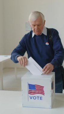 Yaşlı bir adamın oy sandığına oy pusulası yerleştirip Amerika 'daki seçim günü kameraya poz verirken dikey portre çekimi.