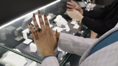 Premium butikte gösterilen zarif mücevherleri deneyen iki kadın müşterinin yakın çekimleri. İsimsiz siyah kadın özel pırıl pırıl yüzüğe hayran.